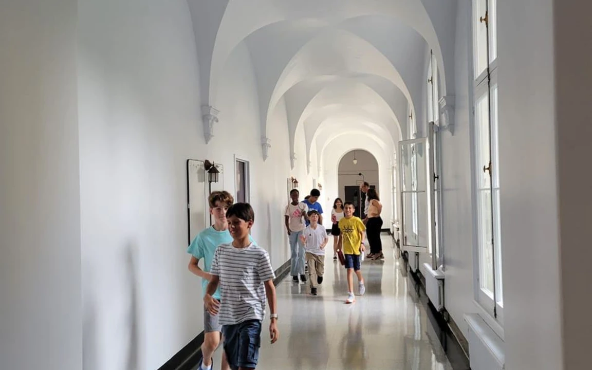  Long couloir lumineux avec arches blanches et fenêtres, sol en pierre lisse menant à une porte au fond.