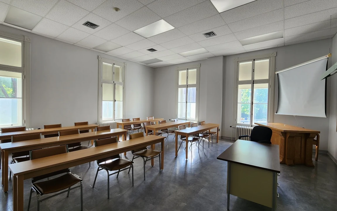 Salle de classe lumineuse avec rangées de bureaux en bois, tableau vert et écran de projection.